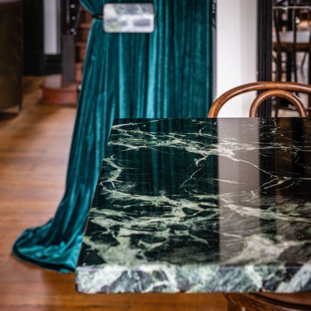 Close-up interior view of a dark green veined marble dining table with a thick polished edge, paired with a curved timber dining chair. A rich teal velvet curtain drapes to the timber floor in the background, with partial architectural and furniture details visible beyond.