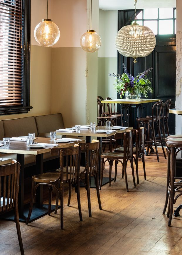 A warmly lit restaurant dining room with closely arranged wooden tables and bentwood chairs, upholstered banquette seating, timber flooring, dark timber wall panelling, and large textured glass pendant lights. A high communal table with floral arrangement sits at the rear, while black-framed glazing and timber blinds add contrast.