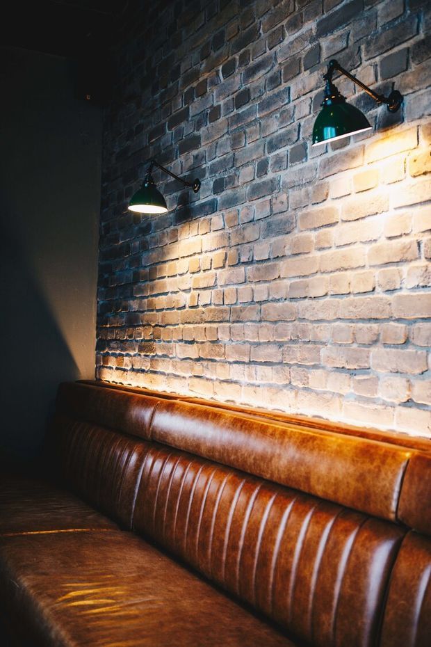 A moody interior corner featuring a built-in brown leather banquette against an exposed brick wall. Two dark green articulated wall sconces cast warm pools of light across the brickwork, creating an atmospheric industrial-style seating area.