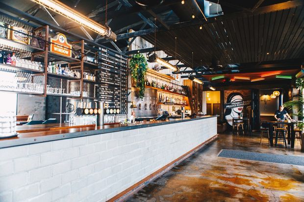 Industrial-style bar and dining venue interior with a long white-painted brick service counter, timber benchtop, exposed dark-painted ceiling structure and ductwork, suspended linear lighting, extensive bottle shelving, and a rear dining area. The space combines raw concrete flooring, black metal detailing, timber accents, and greenery.