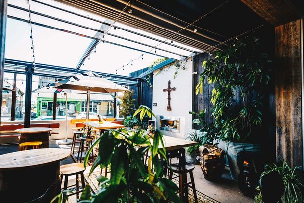 A bright, plant-filled hospitality interior with extensive glass roof and glazing, communal timber tables, stools, pendant string lighting, outdoor-style umbrellas, and a prominent living wall. A crucifix and fireplace-like feature are visible on the white wall, with an urban streetscape seen through the glazing.