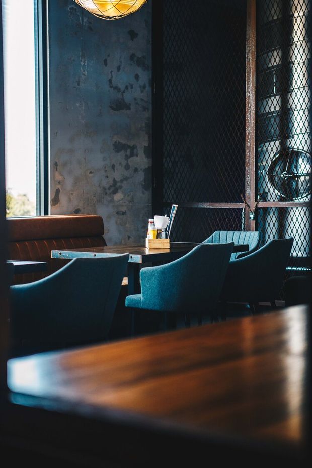 A moody industrial-style dining or lounge area with dark teal upholstered chairs, a long timber table, a leather banquette, distressed blue-grey metal wall panels, and black wire-mesh screening. A warm pendant light and large window add contrast to the dark interior.