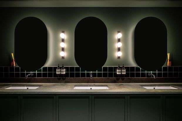 A dark, moody bathroom vanity with three oval mirrors, vertical globe-style wall lights, a long stone benchtop with three integrated basins, dark green panelled cabinetry, and a black tiled splashback.