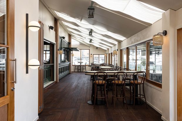 A long, enclosed restaurant dining area with dark timber flooring, timber-framed glazing, and rows of black metal dining tables and chairs. White fabric-like ceiling panels create a softly draped canopy, while contemporary wall sconces and pendant lights provide warm illumination. The space has a refined, inviting hospitality interior with views through the glazing to the surrounding urban setting.