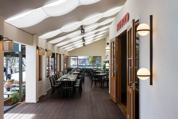 Long, bright restaurant or café dining space with rows of dark tables and chairs, timber-look flooring, large windows and glazed timber-framed doors. A distinctive undulating white fabric-like ceiling treatment runs the length of the room, complemented by decorative pendant and wall lighting. The space opens toward a street-facing glazed frontage and appears inviting and contemporary.
