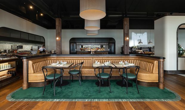 A stylish restaurant or hospitality dining space featuring a curved, upholstered banquette wrapped around a central dining counter, with small tables and dark-framed chairs. The interior combines warm timber flooring and columns, green carpet and upholstery, white walls, black exposed ceiling services, brass-toned details, and large cylindrical pendant lights. A bar and kitchen service area are visible behind the seating.