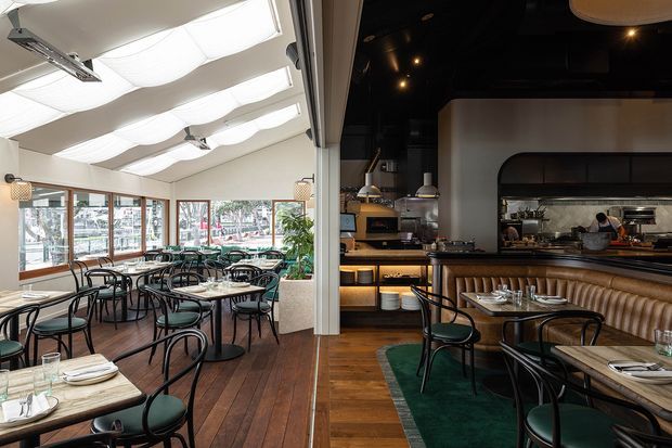 A stylish restaurant interior showing a bright dining room on the left and a darker bar and open kitchen area on the right. The space features timber flooring, marble-topped dining tables, black bentwood-style chairs, upholstered banquette seating, brass-toned pendant lights, and a reflective open-kitchen backsplash.