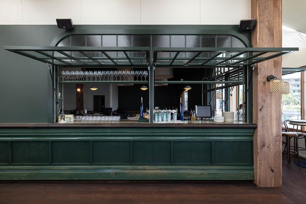 A dark green built-in bar counter with raised decorative paneling, black metal framing, and an overhead glass-and-metal rack displaying wine glasses. The bar is set within a refined commercial interior with timber columns, dark timber flooring, pendant lighting, and visible restaurant seating beyond.
