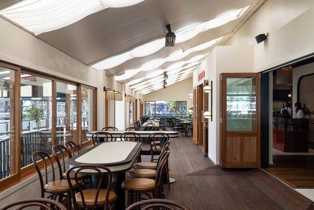 A long, bright restaurant dining area with rows of dark wood tables and curved timber chairs. Full-height glazed doors and windows line the left side, while timber-framed doors and a bar or service area open on the right. The space has dark timber flooring, white walls, and a distinctive undulating fabric-like ceiling treatment with integrated lighting.