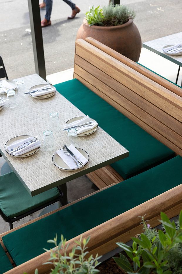 A bright outdoor restaurant dining area viewed from above, with tiled square tables set with plates, cutlery and glasses, surrounded by timber slatted bench seating and deep teal-green cushions. Large terracotta planters with herbs and greenery add a natural element, while dark-framed glazing opens toward the street.