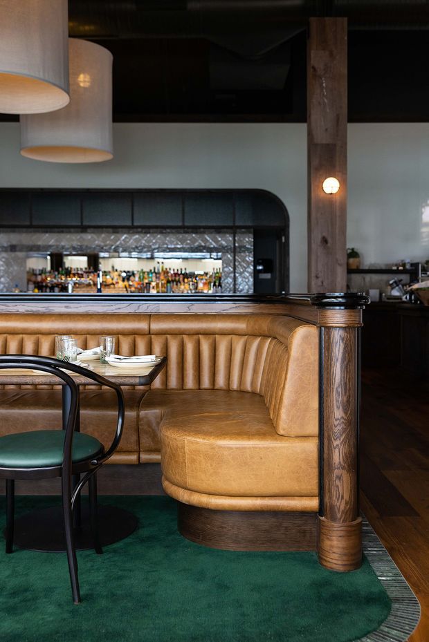 A richly detailed restaurant or bar interior featuring a curved tan leather banquette with vertical channel tufting, dark wood trim and a substantial timber column. A small dining table and dark-framed green upholstered chair sit in the foreground on a deep green carpet. Behind the seating is a back bar with mirrored tile, bottles and shelving, framed by dark arched metalwork. Pendant lights, exposed dark ceiling services and warm timber finishes create an intimate, vintage-inspired atmosphere.