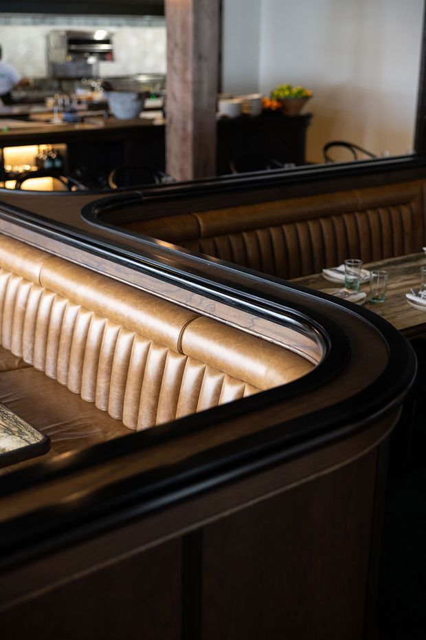 A close-up interior view of a sophisticated dining or lounge booth, featuring deeply channelled tan leather upholstery, dark timber panelling, and glossy black trim. A dining table with glassware is visible behind the curved banquette, while a kitchen or bar area with coffee equipment appears in the background. The image has a warm, refined, restaurant-like atmosphere.