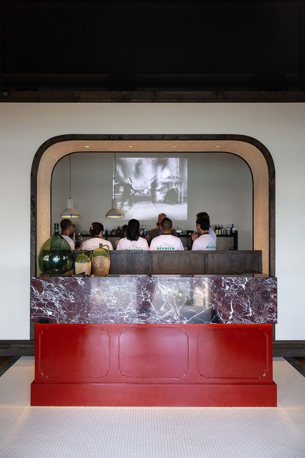 A dramatic hospitality bar or service counter viewed straight on. A deep red paneled base supports a highly veined dark burgundy marble counter, with dark timber cabinetry above. A large rounded-rectangle arched opening frames the bar workspace and a projected black-and-white image on the rear wall. Pendant lights, bottles, vessels, and staff are visible behind the counter. The space has a theatrical, refined, high-contrast character.