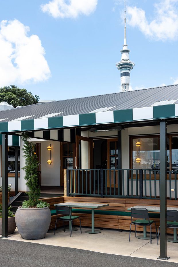 Exterior view of a contemporary hospitality venue with a covered outdoor dining terrace, dark metal framing, timber bench seating, green and white striped awning, warm wall sconces, and large glazed openings. A prominent communications tower rises behind the low pitched metal roof.