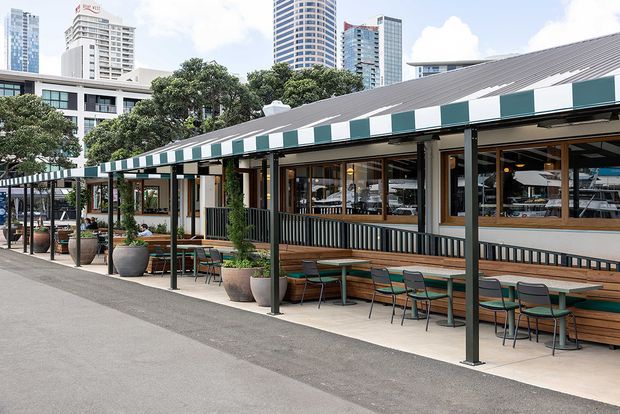 A long, low-rise commercial dining venue with extensive glazed frontage, outdoor tables and chairs, large planters, and a striped green-and-white awning. The building has a pitched metal roof, dark structural posts, timber-framed windows, and a contemporary urban setting with high-rise buildings visible behind it.