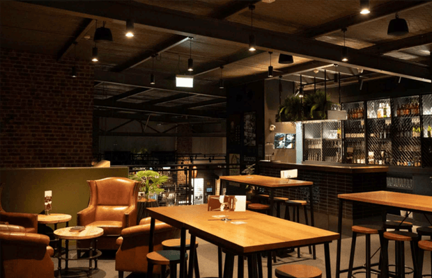 A moody industrial-style hospitality interior featuring a bar and dining area. Warm timber communal tables, black metal bases, leather lounge chairs, exposed red brick walls, dark ceiling beams, pendant lighting, and a backlit bottle display create a sophisticated urban atmosphere.