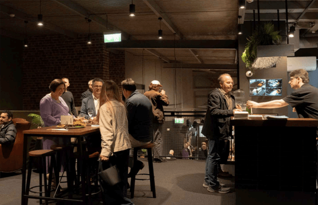A dimly lit contemporary hospitality interior, likely a bar or restaurant, with exposed brick walls, a timber-toned ceiling, pendant lighting, high communal tables, bar stools, and a service counter. Several patrons are gathered around tables and the counter, with a mezzanine or lower-level area visible beyond the railing.