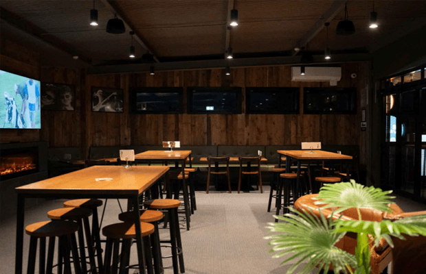 A dimly lit, rustic-modern hospitality interior resembling a bar, café, or restaurant dining area. The space features multiple high timber tables with black metal legs, matching bar stools, dark timber wall panelling, exposed timber ceiling, pendant and track lighting, framed artwork, a wall-mounted screen, and a fireplace along the left wall. Large glazed doors and windows line the right side, while a plant and counter edge appear in the foreground.