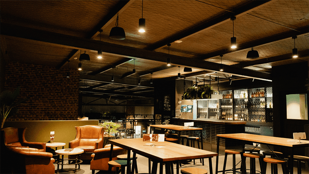 A moody industrial-style bar or restaurant interior with exposed dark brick walls, black steel ceiling beams, suspended pendant lights, timber dining tables, leather lounge chairs, bar stools, and a back bar with illuminated bottle shelving. The space has warm ambient lighting and a casual dining atmosphere.