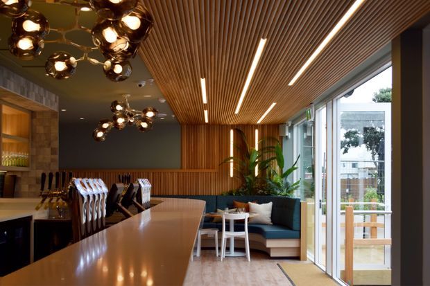 A sophisticated contemporary interior combining a kitchen or bar counter with an adjoining lounge area. The space features a long light stone or composite island, dark cabinetry, warm timber slatted ceiling and wall panelling, sculptural clustered pendant lights, integrated linear lighting, and floor-to-ceiling glazing opening to an outdoor deck or balcony. A compact upholstered seating area with indoor plants sits beside the glazing.