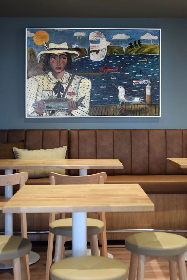 A warmly lit contemporary dining or café interior with timber tables, pale wood chairs, round upholstered stools, and a long brown leather banquette. The walls are painted deep blue-grey and feature a large framed nautical artwork above the seating. The space has a compact, inviting hospitality feel with natural timber and muted upholstery.