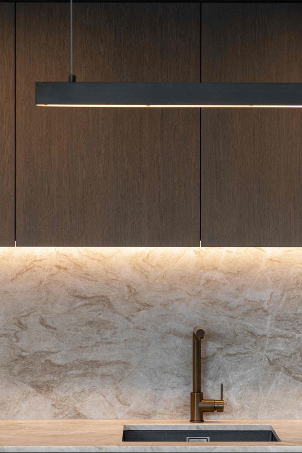 Close-up architectural detail of a refined contemporary kitchen, featuring dark vertically grained timber cabinetry, a long slim black pendant light, a softly backlit beige natural-stone splashback, and a brushed bronze or brass-toned tap above a dark inset sink. A pale timber benchtop and stone foreground surface complete the warm, luxurious composition.