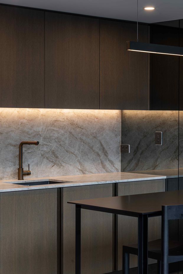 A compact, dark-toned contemporary kitchen with full-height dark wood-grain cabinetry, a pale veined stone backsplash and benchtop, an undermount sink with bronze-toned tapware, under-cabinet lighting, and a black linear pendant above a small dining or breakfast table. A mirrored panel reflects the stone surface and electrical outlets.