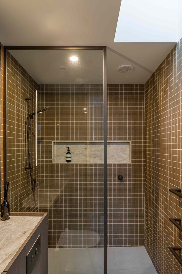 Contemporary bathroom featuring a large walk-in shower enclosed by a frameless glass screen, with brown square wall tiles, pale stone shower niche, dark vanity cabinetry, and a skylight. Brushed or dark metal shower and tapware add contrast to the warm tiled interior.