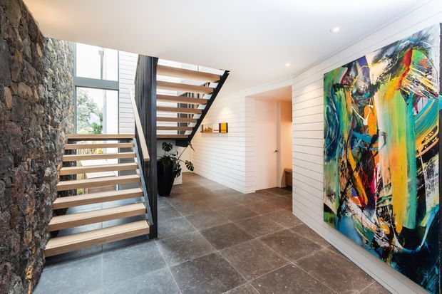 A bright contemporary entry hall with an open-riser timber staircase, dark metal supports, a rugged natural-stone feature wall, white horizontal wall panelling, large grey stone-look floor tiles, and a vivid abstract artwork. Daylight enters through tall glazing beside the stair, creating an airy but dramatic interior.