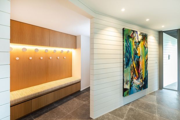 Contemporary entryway with a built-in timber bench and overhead cabinetry, warm integrated lighting, white horizontal wall panelling, dark stone-look flooring, and a colourful abstract artwork. A glazed door and bright adjoining area are visible to the right.