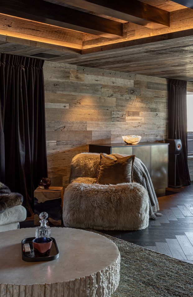 A moody, luxurious living room featuring a fur-covered lounge chair, dark curtains, a low sculptural coffee table, a timber console, and a large wall clad in weathered reclaimed timber. Exposed timber ceiling beams with integrated warm lighting create a dramatic rustic lodge atmosphere, complemented by dark herringbone flooring and a contemporary speaker.