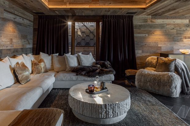 A cozy, moody living room with a large light sectional sofa, plush fur throws and cushions, a sculptural round coffee table, and a textured armchair. The room is enveloped in reclaimed-looking timber wall and ceiling cladding, with dark floor-to-ceiling curtains, warm recessed cove lighting, and a patterned feature window.