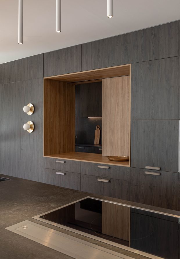 A refined contemporary kitchen featuring full-height dark grey wood-grain cabinetry, integrated drawers, a large dark stone-look island benchtop, and a black glass induction cooktop. A warm timber-lined recessed display niche contrasts with the darker cabinetry, while minimalist cylindrical pendant lights and sculptural wall sconces provide understated illumination.