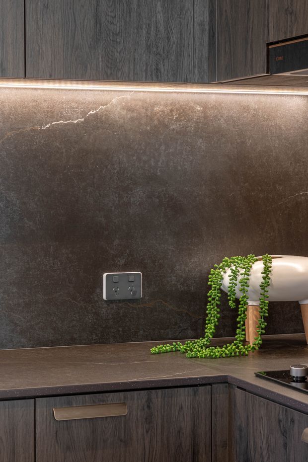 Close-up view of a dark, contemporary kitchen corner featuring charcoal wood-grain cabinetry, a dark mottled stone-look splashback and benchtop, integrated warm LED strip lighting, a power outlet, and a black cooktop. A trailing plant and small appliance add decorative detail.