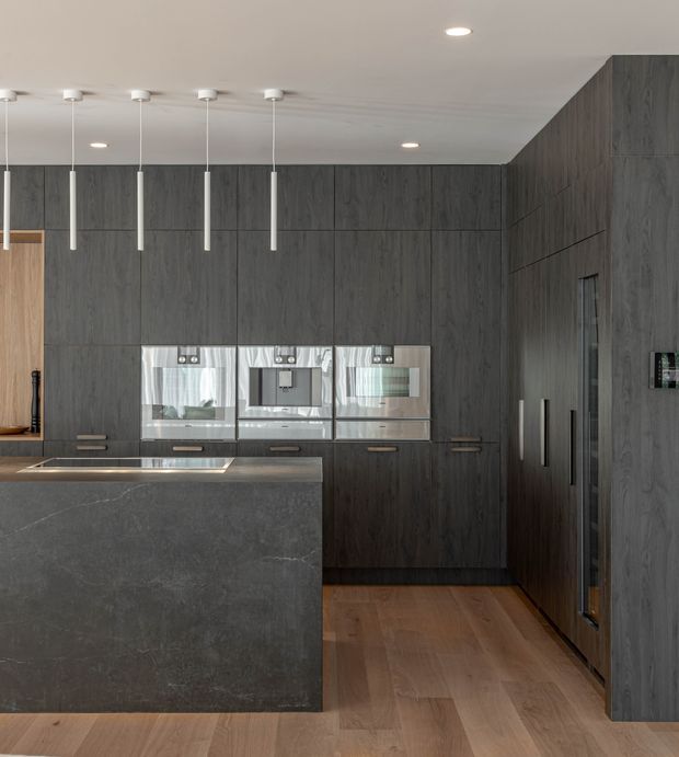 Contemporary kitchen featuring extensive dark wood-grain full-height cabinetry, integrated stainless-steel ovens, a large charcoal stone island with waterfall edge, timber flooring, and slim white pendant lights beneath a smooth white ceiling.