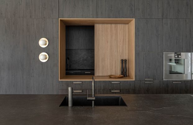 A close-up view of a sleek contemporary kitchen featuring dark vertically grained wall cabinetry and a dark stone-look benchtop with an integrated double-bowl sink and minimalist tapware. A warm timber-framed open cabinet with sliding timber doors provides contrast, while built-in stainless-steel appliances are visible to the right. Two small round wall lights add subtle illumination.