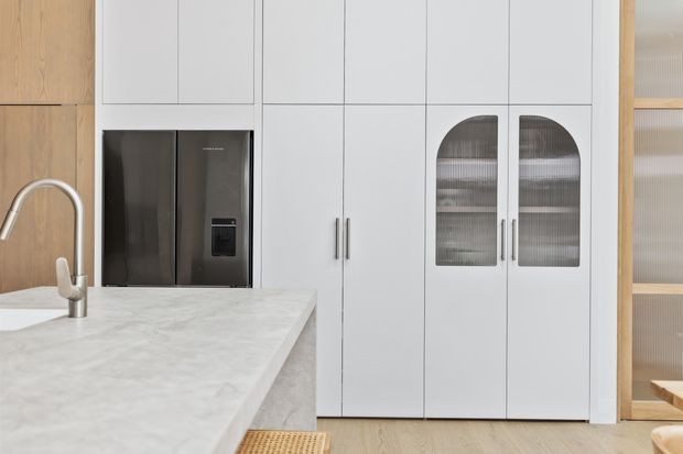 Bright contemporary kitchen with a large pale stone island benchtop, integrated dark refrigerator, white full-height cabinetry, and distinctive arched reeded-glass cabinet doors. Light timber cabinetry and flooring add warmth, while brushed metal handles and a curved stainless tap provide understated contrast.