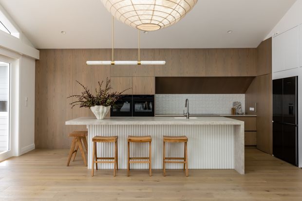 A bright, open-plan contemporary kitchen centred on a large island with a vertically fluted white front and pale stone benchtop. Warm timber cabinetry and matching timber flooring create a natural backdrop, with integrated black appliances, a pale textured tile splashback, timber bar stools, and a sculptural woven pendant light.