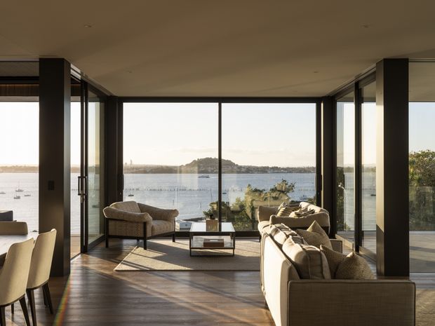 A refined contemporary living room with floor-to-ceiling glazing overlooking a calm waterfront and distant headland. The room features dark timber flooring, slim black-framed glass doors and windows, neutral upholstered sofas and armchairs, a low coffee table, and a dining area at the left. Warm natural light creates a relaxed, sophisticated atmosphere.