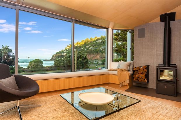 A warm, refined living room with expansive floor-to-ceiling glazing overlooking a leafy coastal landscape and water. The room features a built-in timber window seat and cabinetry, a large glass coffee table, a sculptural dark lounge chair, a textured neutral rug, and a freestanding black wood-burning stove with stacked firewood. The ceiling and window reveals are timber-toned, creating a calm connection to the surrounding landscape.