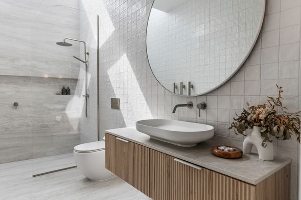 Bright contemporary bathroom featuring a walk-in glass shower with large pale stone-look wall panels, a wall-hung fluted timber vanity with a light stone benchtop, white vessel basin, wall-mounted brushed metal tapware, and a large round mirror. Small-format white tiles surround the vanity, while neutral finishes and dried botanical styling create a calm, refined atmosphere.