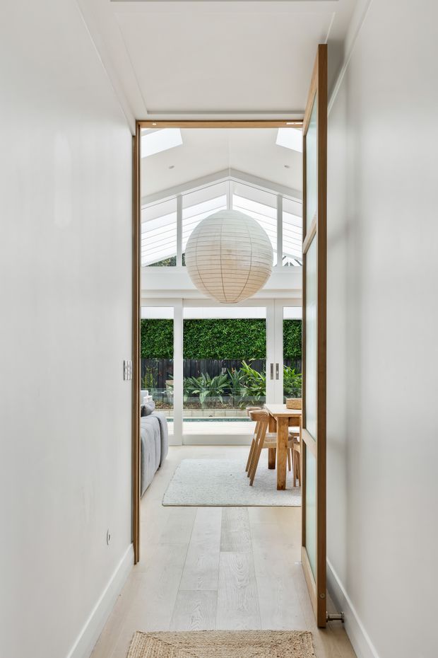 A bright interior view through a timber-framed doorway into an open-plan living and dining area. The room features pale timber flooring, white walls and ceiling, expansive glazing, a large woven pendant light, timber dining furniture, and a planted outdoor courtyard visible beyond. A high glazed roof structure creates an airy, light-filled atmosphere.