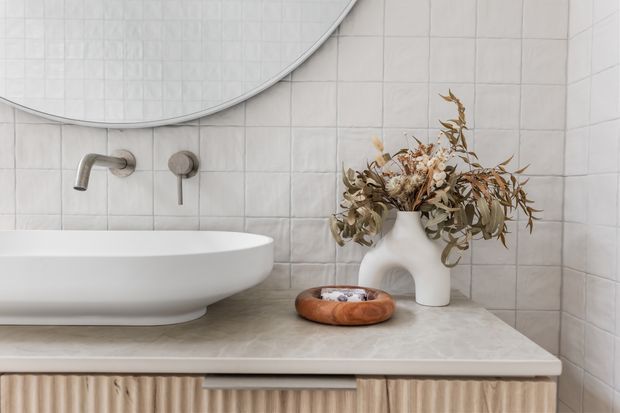 Close-up of a refined contemporary bathroom vanity with a white vessel basin on a pale stone benchtop, brushed metal wall-mounted tapware, softly textured light tiles, and a large rounded mirror. Fluted light-wood cabinetry is visible below, complemented by sculptural white and terracotta accessories and dried botanical stems.
