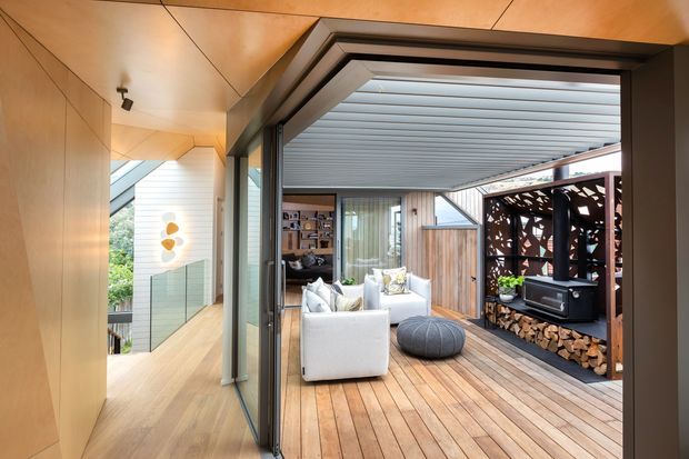 A contemporary indoor-outdoor living area viewed from a timber-lined interior hallway. Fully open glazed doors lead to a covered timber deck furnished with white lounge chairs and a round ottoman. A built-in outdoor fireplace with stacked firewood sits against a dark decorative screen, while timber cladding, warm wood surfaces, and extensive glazing create a seamless connection between the interior and terrace.