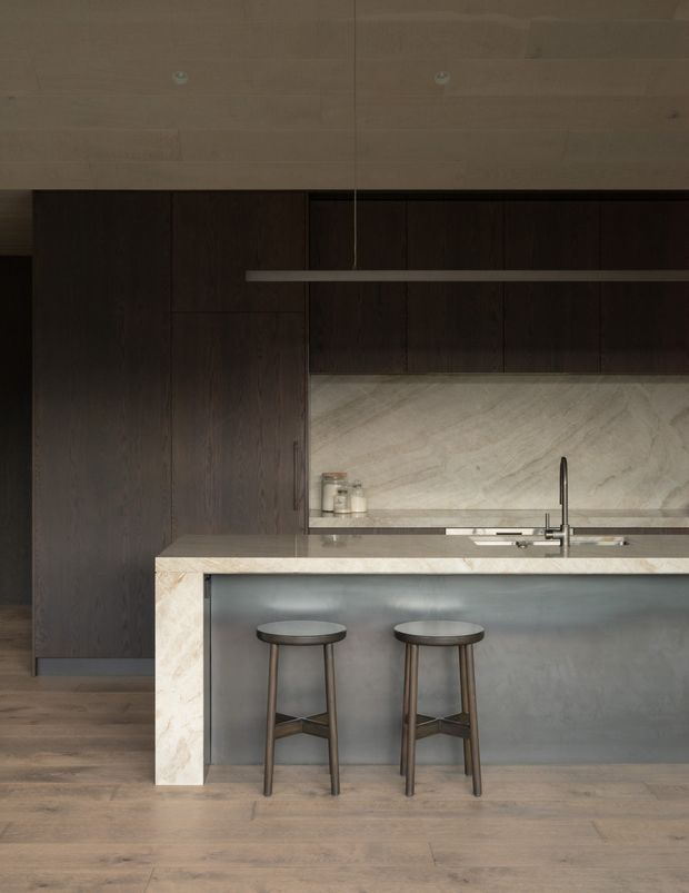 A moody, refined contemporary kitchen centered on a large island with seating for two. The kitchen features dark timber cabinetry, a pale stone island benchtop and waterfall end panel, a grey integrated island face, matching pale stone backsplash, timber flooring, and a slim linear pendant light. A concealed or integrated rangehood and sink with a tall tap are visible along the rear counter.