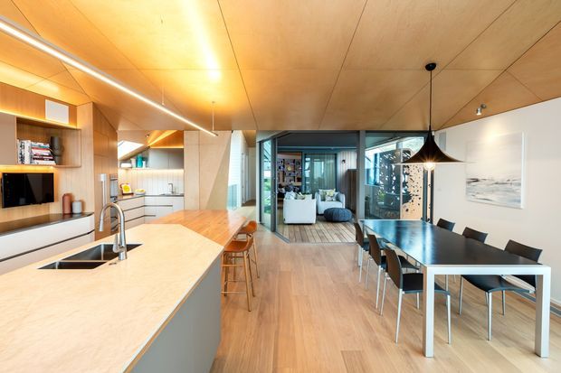 Open-plan contemporary kitchen, dining and living area with extensive warm timber wall and ceiling panelling. The kitchen features a large pale stone island with integrated sink, light cabinetry and timber open shelving. A black pendant hangs above a dark dining table with modern chairs, while glazed doors open toward a bright living room beyond. The space is warm, refined and naturally lit.