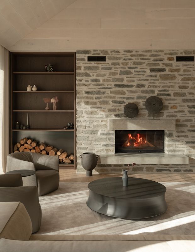 Warm, refined living room centred on a stone masonry fireplace with a built-in firebox and thick pale stone mantel. A dark timber built-in shelving and firewood storage unit occupies the left side, while curved upholstered lounge chairs and a low rounded dark coffee table sit on a large neutral rug. The ceiling is clad in pale timber boards, and sunlight creates strong diagonal shadows across the room.