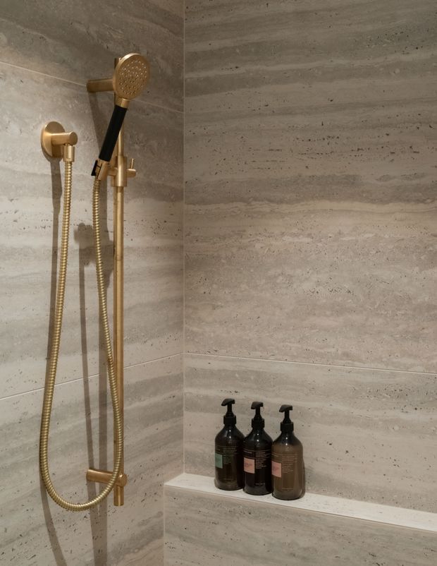 Close-up view of a contemporary shower enclosure clad in large-format horizontally veined grey-beige stone or porcelain panels. A brushed brass handheld shower set with wall-mounted rail and flexible hoses is mounted on the left wall, while three dark pump bottles sit on a recessed corner shelf. The bathroom has a warm, refined, spa-like appearance with minimal detailing.