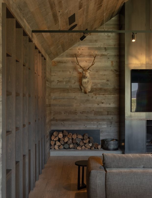 A moody, sophisticated living room with a timber-lined sloped ceiling and reclaimed-looking horizontal timber feature wall. Full-height dark timber built-in cabinetry lines the left side, while a fireplace with stacked firewood and a mounted deer head forms the focal point. A grey upholstered sofa, black side table, dark metal fireplace surround, and wall-mounted television occupy the foreground and right side. Warm directional track lighting enhances the rustic, lodge-like atmosphere.