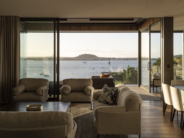 A refined living room with expansive floor-to-ceiling glazed sliding doors overlooking a calm bay and a small island. The space features several cream boucle upholstered armchairs and sofas, a dark coffee table, timber flooring, full-height curtains, and warm timber-lined ceiling detailing around the opening. A dining area is visible to the right, while the waterfront outlook dominates the room.